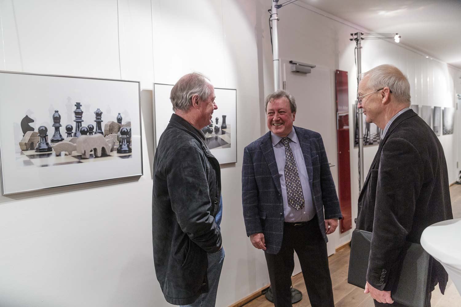 Drei Herren unterhalten sich lachend in der Ausstellung, an der Wand eine Fotografie mit Schachfiguren und Holztieren auf dem Brett.