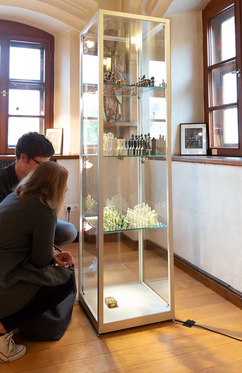 Zwei Besucher hocken vor einer beleuchteten Glasvitrine und betrachten mehrere Schachspiele auf den Glasböden